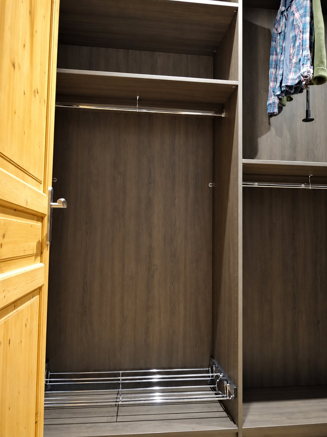 Intérieur d’un dressing en bois foncé avec étagères, tringles de penderie, panier coulissant chromé et porte en pin ouverte.