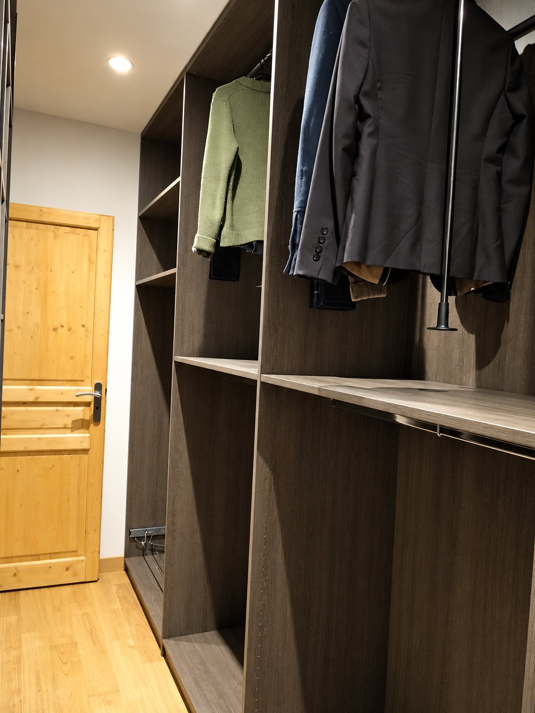 Dressing sur mesure aux étagères en bois sombre, avec une veste verte et des blazers suspendus, près d’une porte en bois clair.